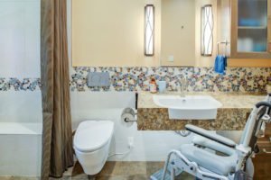 A universal design bathroom with a tiled accent has a roll-under stone vanity, wall-mounted toilet, and curbless shower with a bench.