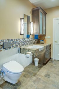 A universal design bathroom with a tiled accent has a roll-under stone vanity and a wall-mounted toilet with a flush plate.