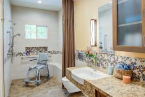 A universal design bathroom with a tiled accent has a roll-under stone vanity, wall-mounted toilet, and curbless shower with a shower wheelchair in it.