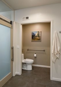 A contemporary universal design bathroom has an extra wide doorway leading to the toilet so it can be accessed by someone in a wheelchair. Brushed metal railings are on the wall to the left of and behind the toilet.